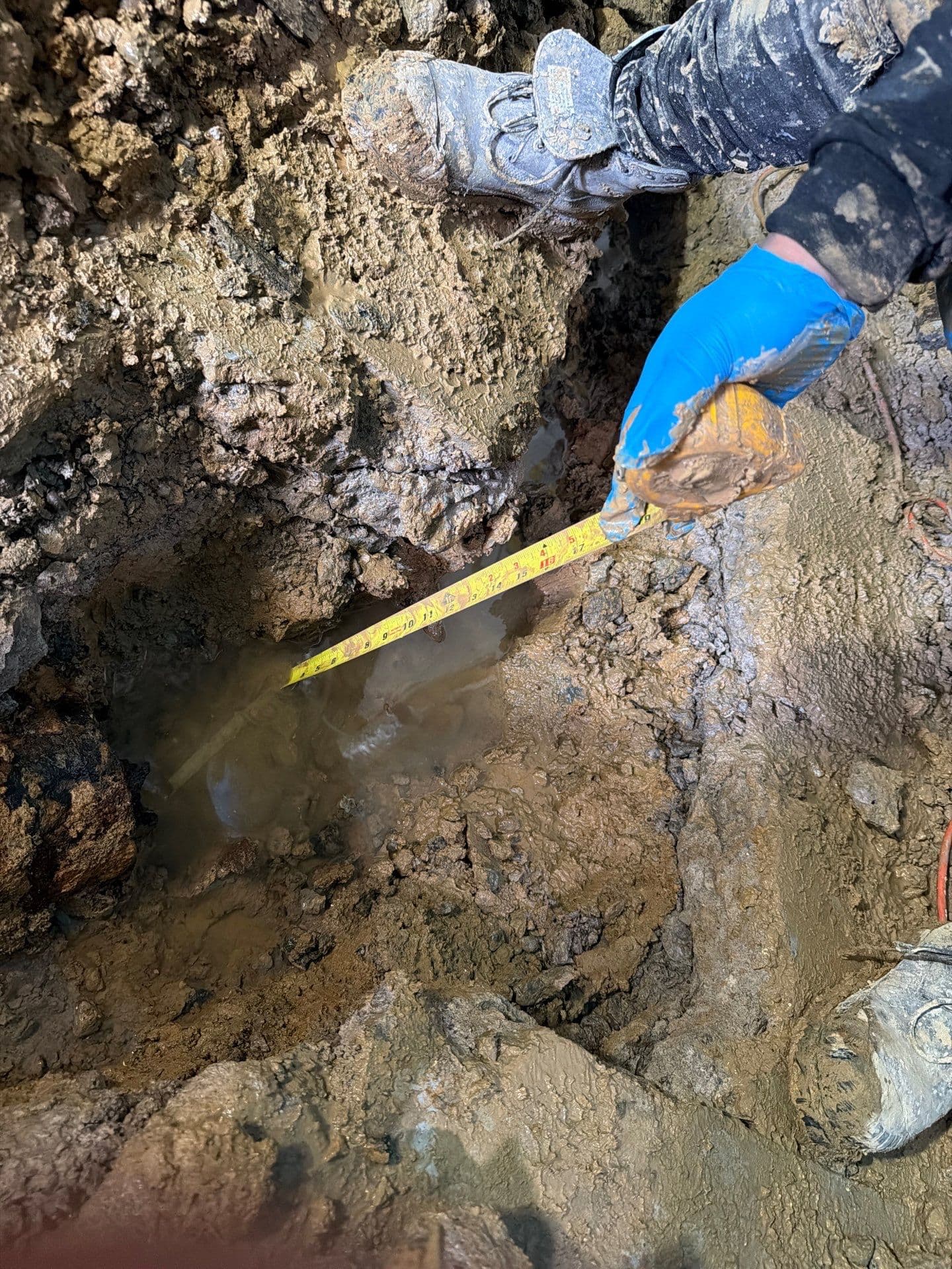 Person in blue gloves measuring a water-filled muddy hole with a yellow tape measure.