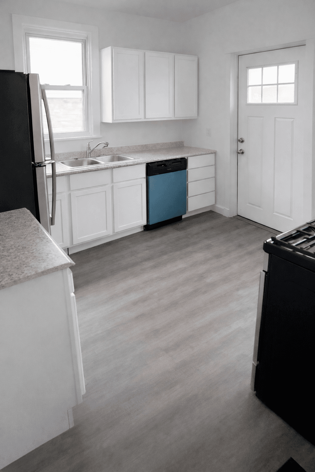Bright kitchen with white cabinets, grey floors, a blue dishwasher, and a white exterior door.