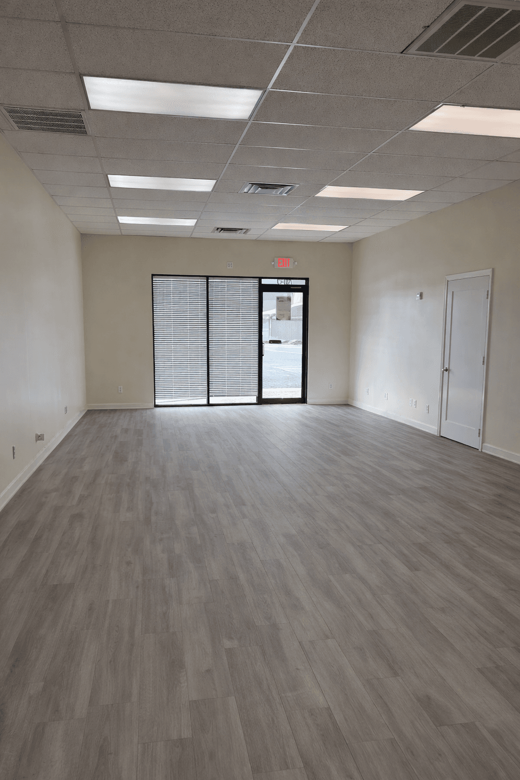 Empty room featuring grey wood-look flooring, white walls, drop ceiling, and glass exit door.