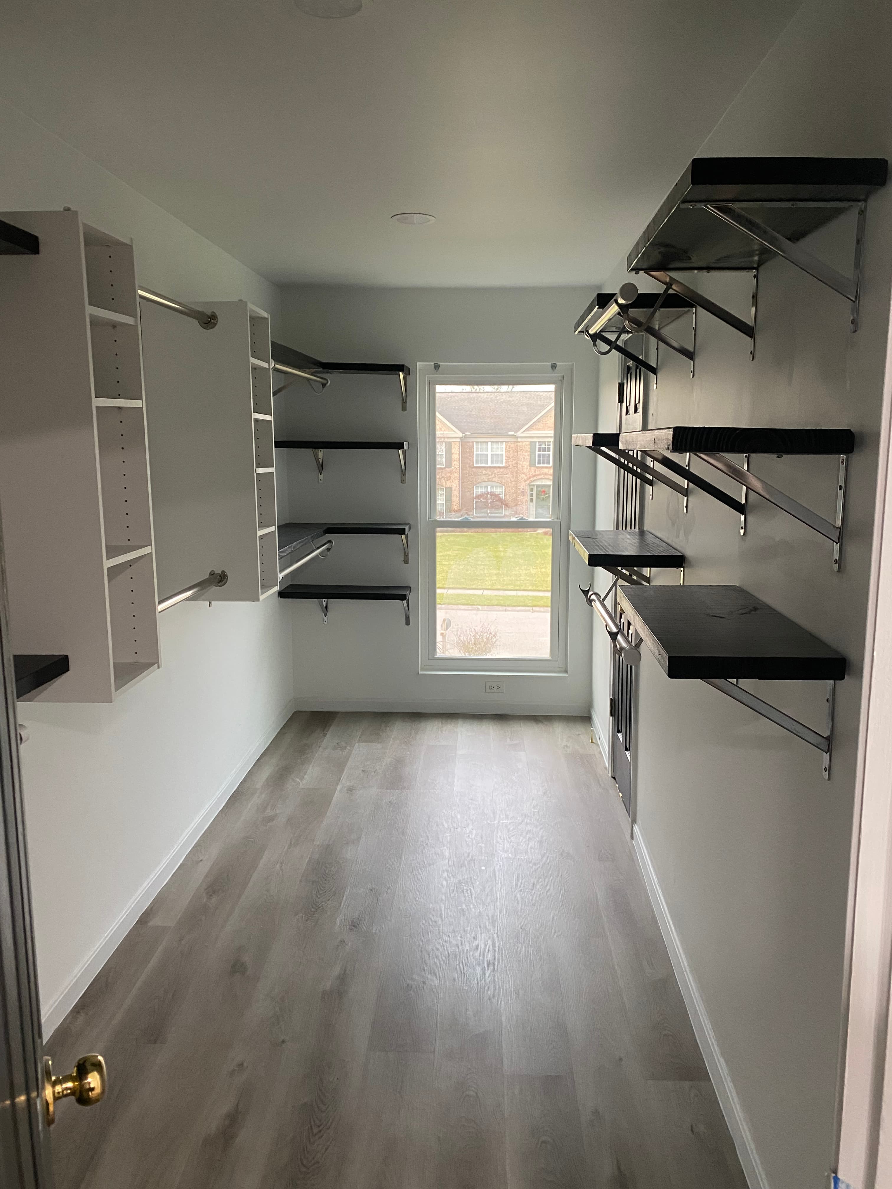 Empty walk-in closet with white walls, wood flooring, mixed shelving, and a central window.